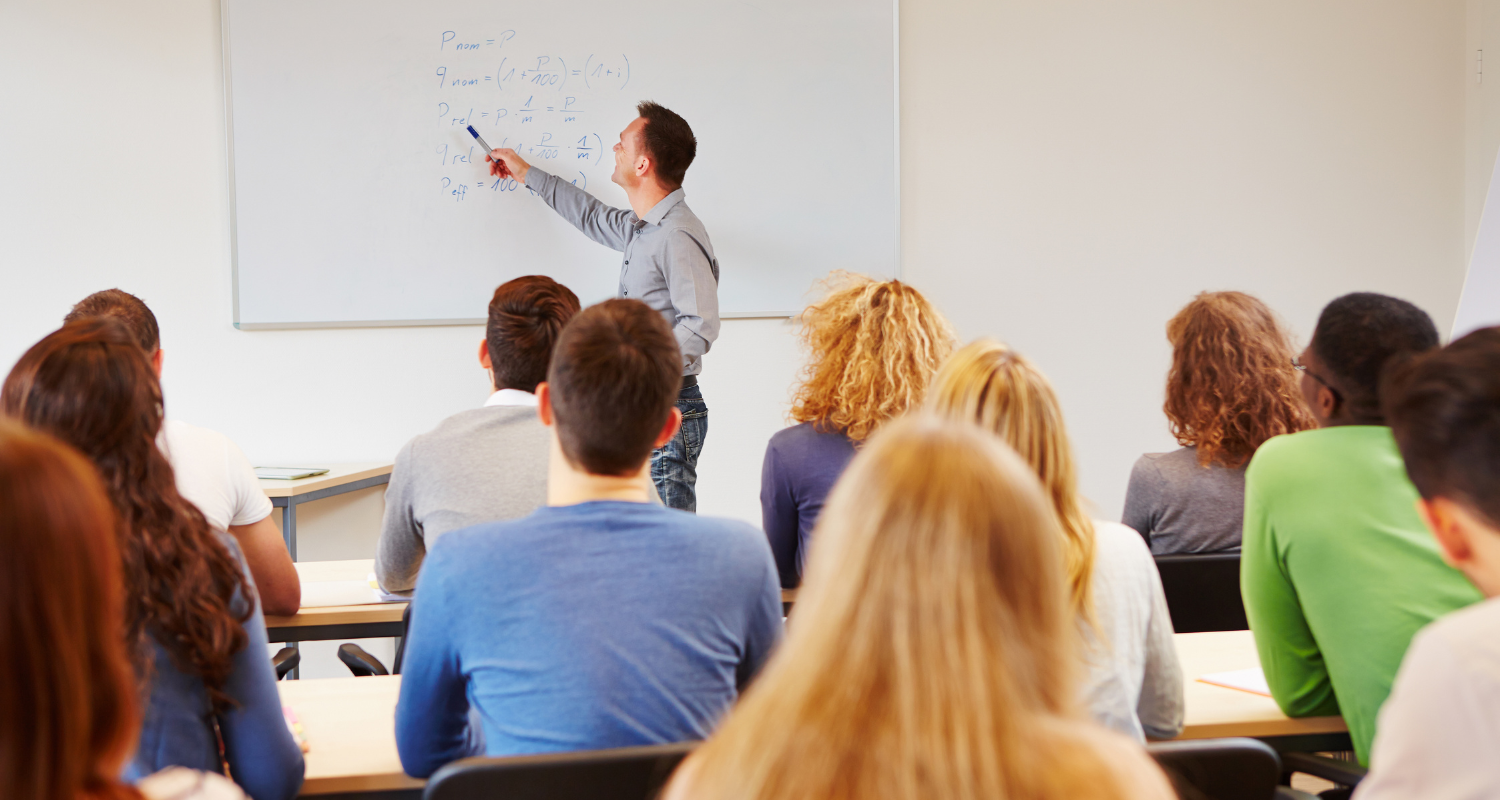 The teacher points to the blackboard, behind him are adults