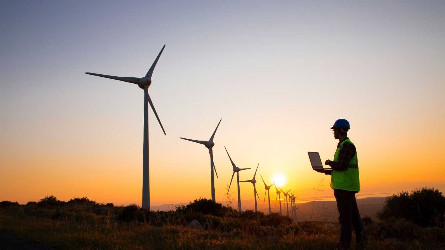 A worker in a helmet and a reflective vest with a laptop looking at windmills